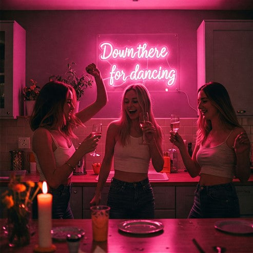 Three women enjoying a vibrant kitchen illuminated by a pink neon sign that reads 'Down there for dancing,' showcasing how neon signs can brighten up and add personality to your space