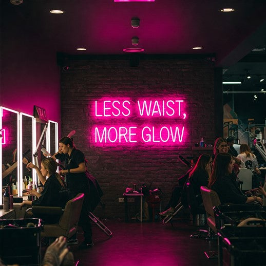 Vibrant pink LED neon sign reading 'Less Waist, More Glow' lighting up a stylish salon, creating an inspiring and modern atmosphere for beauty and self-care spaces.